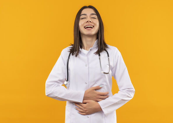 人年轻的女医生穿着白大褂 脖子上戴着听诊器 站在橘色的墙上笑着摸着肚子听诊器笑站