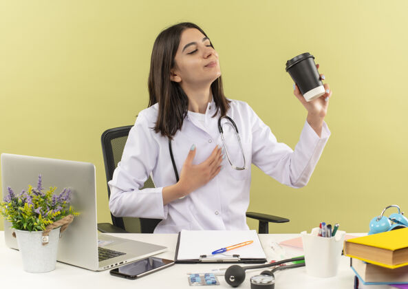 工作年轻的女医生穿着白大褂 脖子上戴着听诊器 手里拿着咖啡杯 坐在桌子旁 笔记本电脑放在浅色的墙上坐着医院抱着