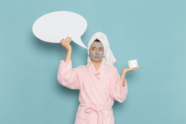 浴袍正面图年轻女性穿着粉色浴袍淋浴后手持巨大的白色标志在蓝色墙壁上美容水霜自理淋浴浴室成人人自理