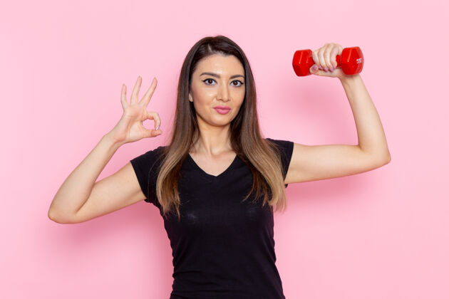 哑铃正面图年轻女性手持浅粉色墙上的红色哑铃运动员运动锻炼健康锻炼女性举行轻