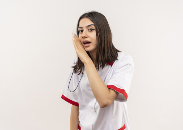 女人年轻的女医生穿着白大褂 脖子上戴着听诊器 手靠嘴站在白墙上讲着一个秘密医生人秘密