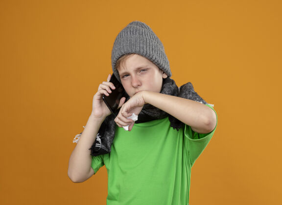 手机生病的小男孩穿着绿色t恤 戴着暖和的围巾和帽子 不舒服地用手机说话 用纸巾擦鼻子站在橙色的墙上穿疾病橙色