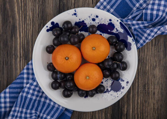 盘子顶视图：白色盘子上的橘子和李子 木制背景上的蓝色格子毛巾毛巾白色水果色