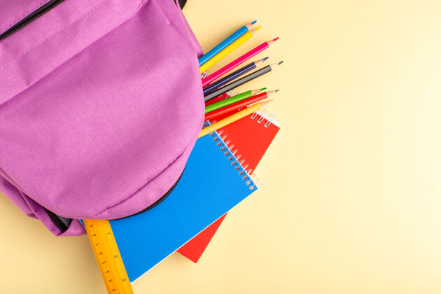 浅黄色顶视图彩色铅笔与抄写本和浅黄色墙上的紫色书包学校毛毡笔铅笔本记事本铅笔学校包