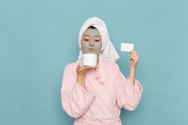 医疗正面图年轻女性穿着粉色浴袍淋浴后拿着奶油和卡片在蓝色墙壁上美容水霜自理淋浴浴室面罩保持后面