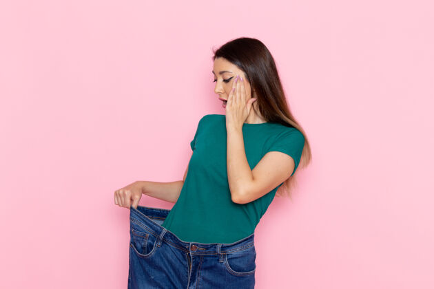 腰部正面图身着绿色t恤的年轻女性在浅粉色墙壁上检查腰部腰部运动锻炼美丽苗条运动员女性运动观点运动