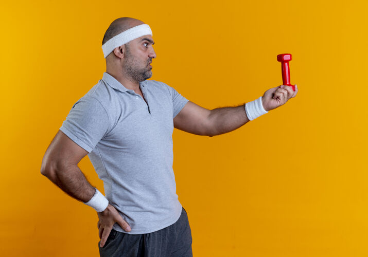 教练成熟的运动型男人戴着头巾 举着哑铃 严肃的脸站在橙色的墙上看着它看严肃成熟