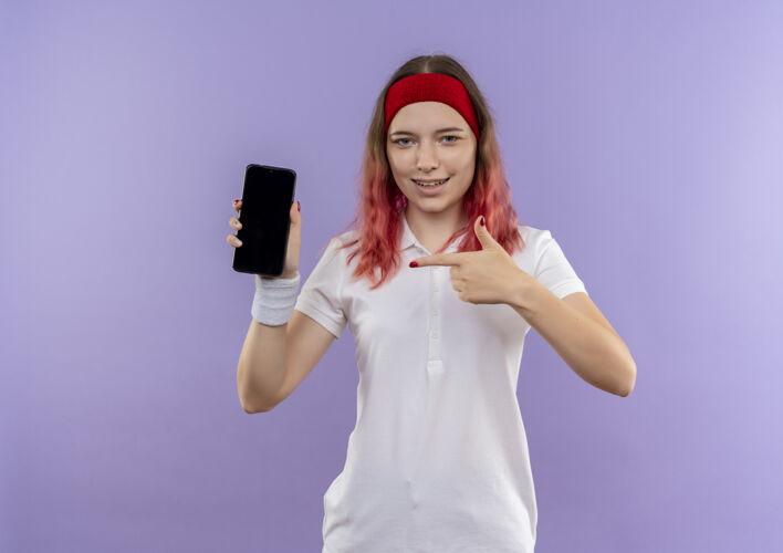 运动装年轻的运动型女人站在紫色的墙上 用手指指着智能手机 带着自信的微笑表演移动微笑