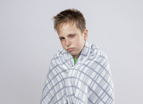 男孩生病的小男孩穿着绿色t恤 裹着毯子 站在白色的墙上 感觉不舒服 看起来很不开心 很不舒服小毯子站着