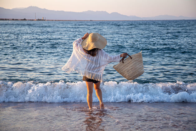 海岸一个戴着大帽子和柳条袋的女孩沿着海边散步暑假概念海浪日落女人