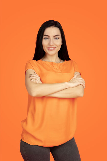 成功橙色工作室背景上的白种人年轻女子肖像穿着衬衫的漂亮黑发女性模特人类情感的概念 面部表情 销售 广告广告空间站着交叉手行政面部女性