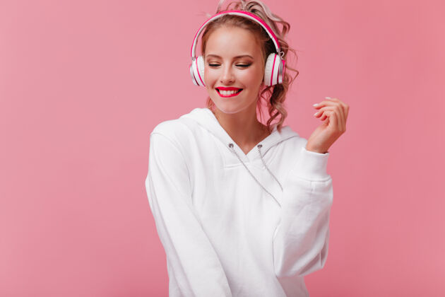 人年轻女子摆姿势 通过她的粉红色耳机听音乐微笑多彩金发
