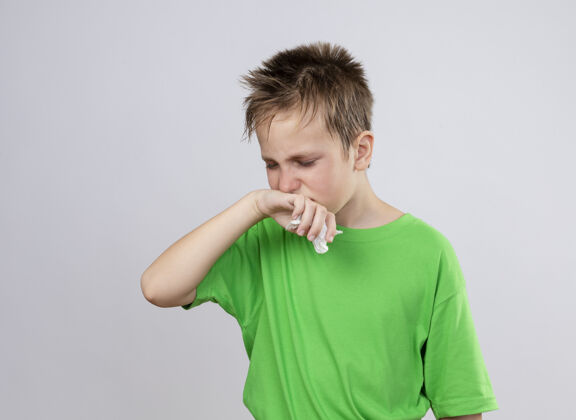痛苦生病的小男孩穿着绿色t恤 站在白墙上流鼻涕和流感 感觉不舒服感觉鼻子男孩