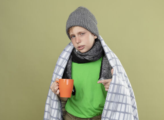 穿上生病的小男孩穿着绿色t恤 戴着暖和的围巾 帽子裹着毯子 手里拿着一杯热茶 手指指着它站在明亮的墙上围巾包着温暖