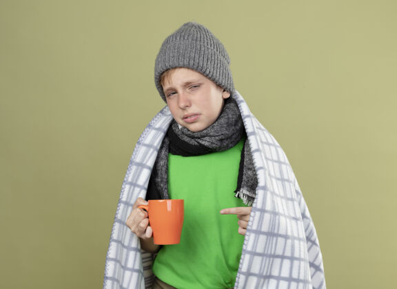 毯子生病的小男孩穿着绿色t恤 戴着暖和的围巾 帽子裹在毯子里 手里拿着一杯热茶 手指指着它 站在明亮的墙上 不开心 生病光手指小