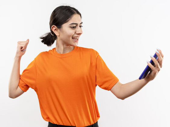 抱着身穿橘色t恤的年轻女孩手持智能手机握紧拳头站在白色的墙壁上快乐而兴奋橙色站着拳头
