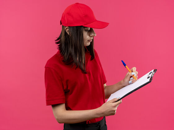 写字穿着红色马球衫 戴着帽子 手里拿着写字板 手里拿着钢笔 站在粉红色的墙上写着一些看起来很自信的东西的年轻送货女孩马球什么的拿着