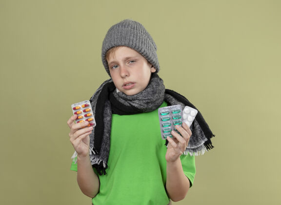 温暖生病的小男孩穿着绿色t恤 戴着暖和的围巾和帽子 感觉不舒服 拿着药片 不高兴 生病地站在光墙上感觉站不开心
