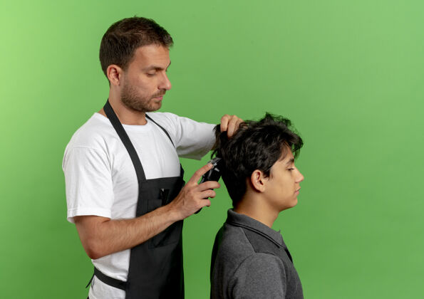男人穿着围裙的理发师站在绿色的墙上 用满意的顾客的剪子剪头发姿势满意修剪