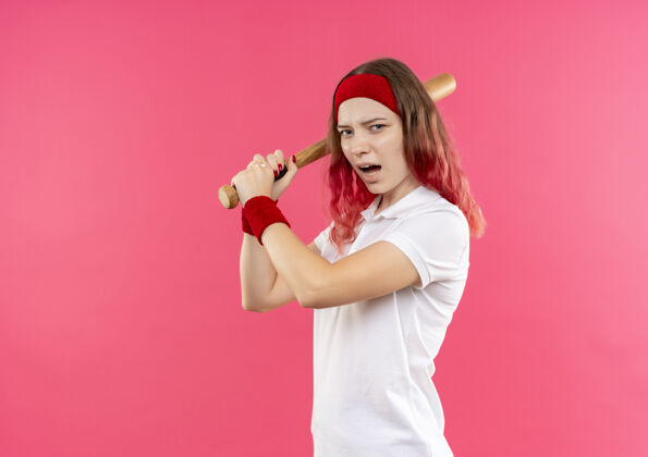 市民戴着头巾的年轻运动女郎挥舞着球棒 站在粉红色的墙上打棒球站立头带运动