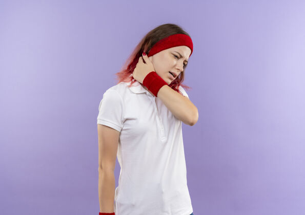 疼痛一个年轻的运动型女人站在紫色的墙上摸着她的脖子 感觉很痛人感觉触摸