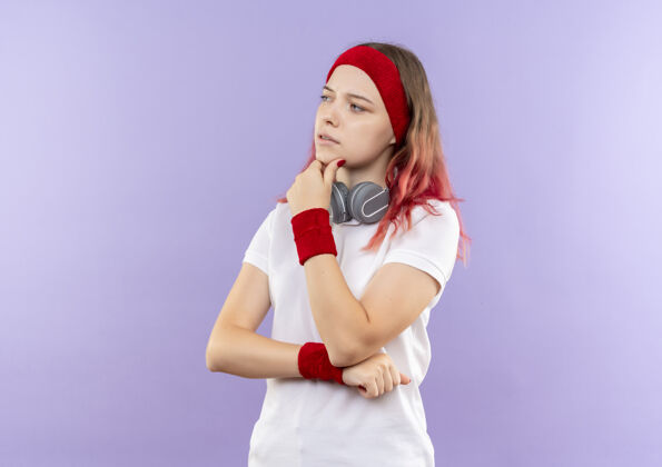 人戴着耳机 手放在下巴上 站在紫色墙壁上思考的年轻运动女性姿势站运动