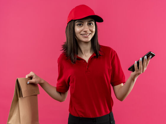 抱年轻的送货女孩穿着红色马球衫和帽子拿着纸包和智能手机微笑友好地站在粉红色的墙上帽子马球站