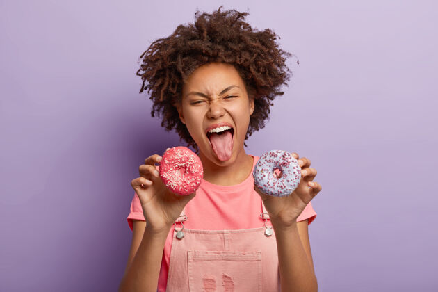 卡路里疯狂情感的黑皮肤少女露出舌头 拿着两个美味的甜甜圈甜点青少年满意