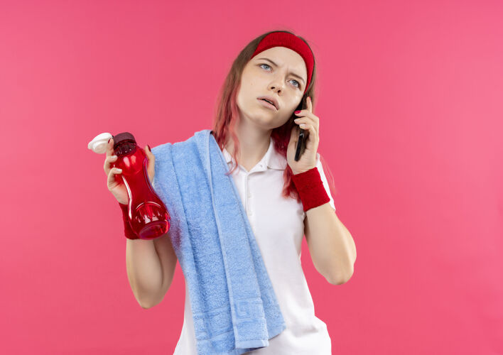 运动戴着头巾 肩上扛着毛巾的运动型年轻女子一边拿着一瓶水 一边用手机聊天 一边严肃地站在粉红色的墙上瓶子而人