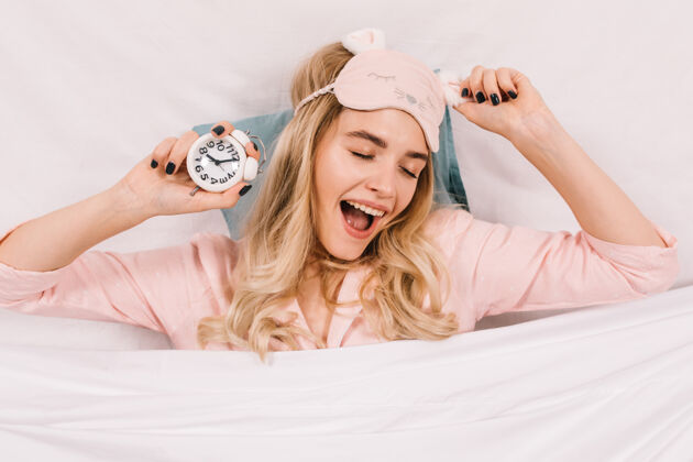 年轻人穿着粉色睡眠面具的优雅年轻女子与时钟合影成人室内早起
