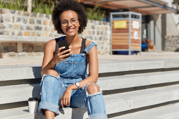 女士皮肤黝黑的女性在手机上发短信 在社交网络上聊天 穿着破烂的工作服 坐在楼梯上 享受一次性咖啡 在街上消遣无忧无虑的青少年和设备饮料短信牙套