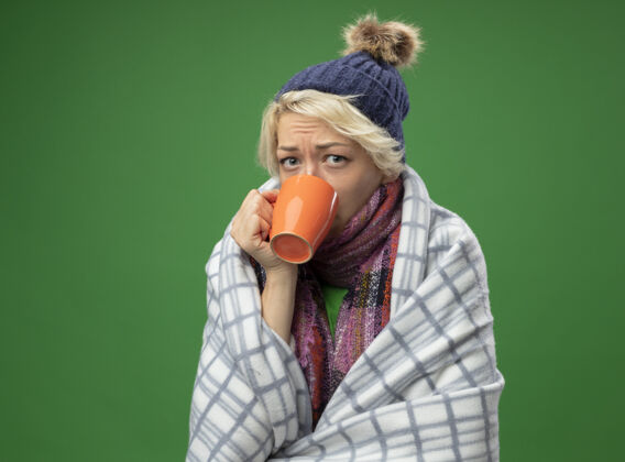 茶生病的不健康的妇女 短头发 戴着暖和的围巾和帽子 感觉不舒服 裹着毯子 发烧 站在绿色的背景上喝着热茶感觉包裹不适