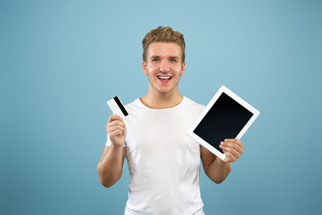 人以蓝色工作室为背景的白人青年半身像穿着衬衫的漂亮男模人类情感的概念 面部表情 销售 广告显示平板电脑屏幕和支付卡工作男性表情