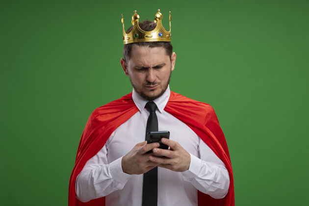 站超级英雄商人穿着红色斗篷戴着皇冠用智能手机看着它严肃的脸站在绿色的背景下商人斗篷严肃