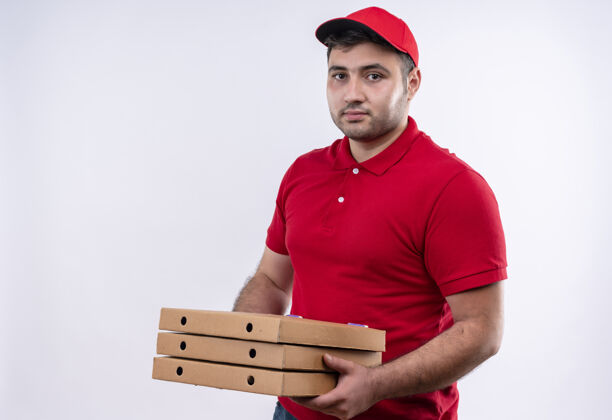 人年轻的送货员身穿红色制服 头戴鸭舌帽 手里拿着披萨盒 站在白色的墙上 脸上带着自信的微笑帽子拿着制服