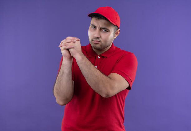 使年轻的送货员身穿红色制服 戴着帽子 手拉手 严肃的脸站在紫色的墙上人脸认真