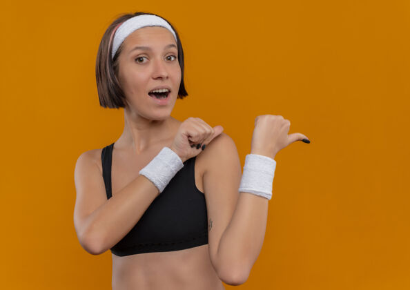 背部穿着运动服的年轻健身女士 头上戴着积极的头带 高兴地向后指着 大拇指站在橙色的墙上运动装运动头带