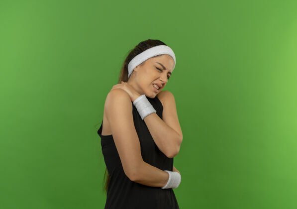 健身穿着运动服的年轻健身女士戴着头带 看起来不舒服 她站在绿色的墙上 摸了摸肩膀 感到疼痛人头带姿势