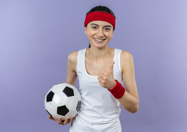 人身穿运动服 头箍 手持足球 微笑自信 竖起大拇指的年轻健身女士站在紫色的墙上微笑公民姿势