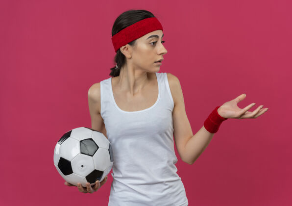 健康身穿运动服的年轻健身女 头戴足球带 手臂伸出 站在粉红色的墙上 看着一边人足球运动