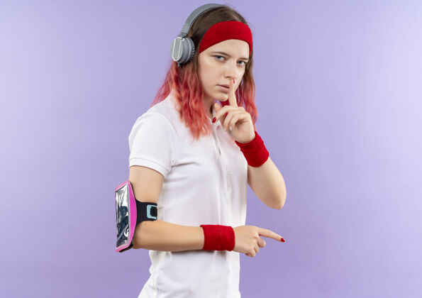 公民戴着耳机戴着头带的年轻运动女性站在紫色的墙上做着手指放在嘴唇上的沉默手势嘴唇耳机沉默