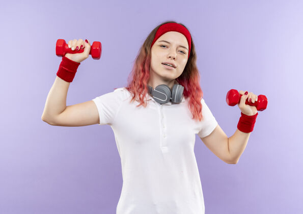 自信年轻的运动型女子举着两个哑铃 自信地微笑着站在紫色的墙上练习女人人运动员