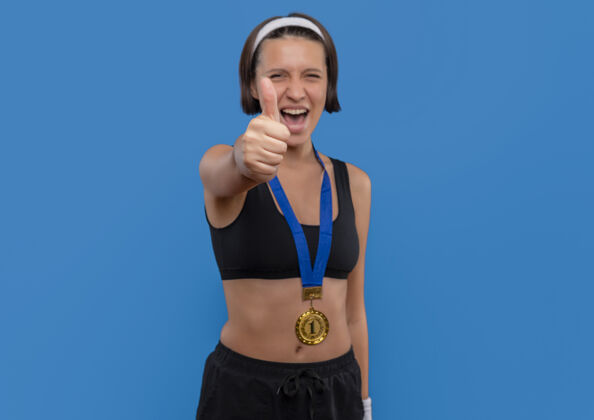 市民一位身穿运动服 脖子上挂着金牌的年轻女子竖起大拇指 为自己的成功而欢欣鼓舞 她站在蓝色的墙上 快乐而兴奋人人运动员