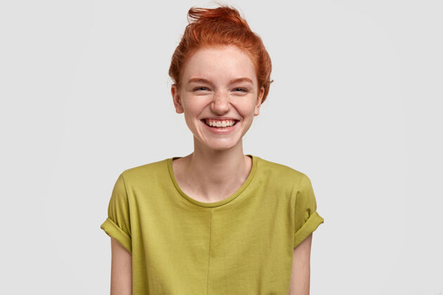 牙关可爱的红发女孩 表情积极 笑着看搞笑的电视节目 享受周末 穿着绿色t恤 皮肤有雀斑 隔着白墙 被滑稽的想法逗乐了感情情绪结