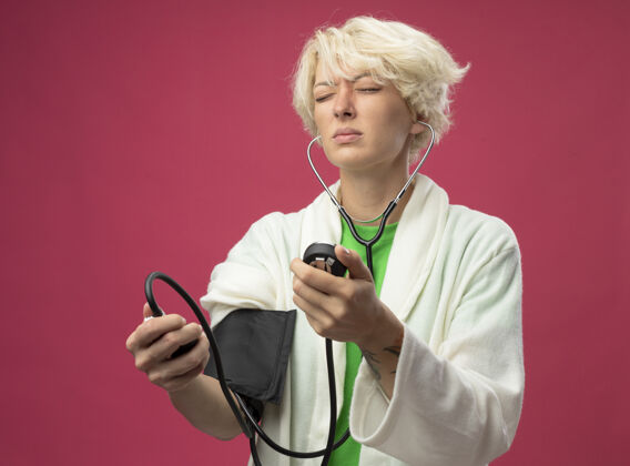 不适有病的不健康妇女 短发 用听诊器测量血压 站在粉色背景上感觉不舒服听诊器不健康测量