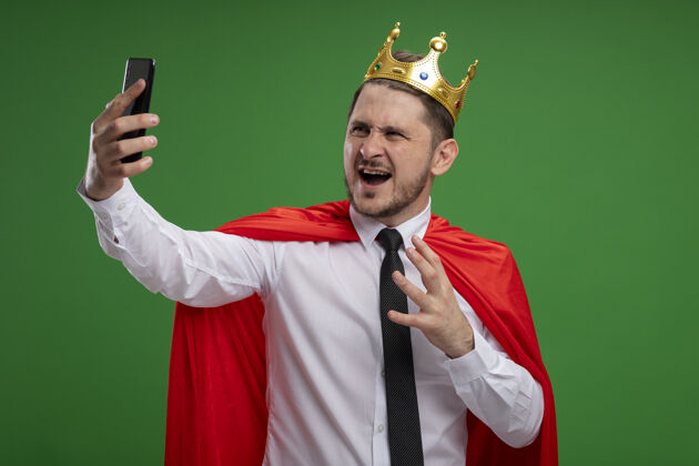 愤怒穿红斗篷戴皇冠自拍的超级英雄商人用智能手机疯狂愤怒地站在绿色背景下皇冠商务人士移动