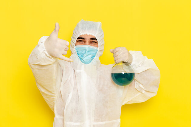 正面正面图穿着特殊防护服的男科学工作者拿着装有蓝色溶液的烧瓶摆在黄色的桌子上医疗医生特殊