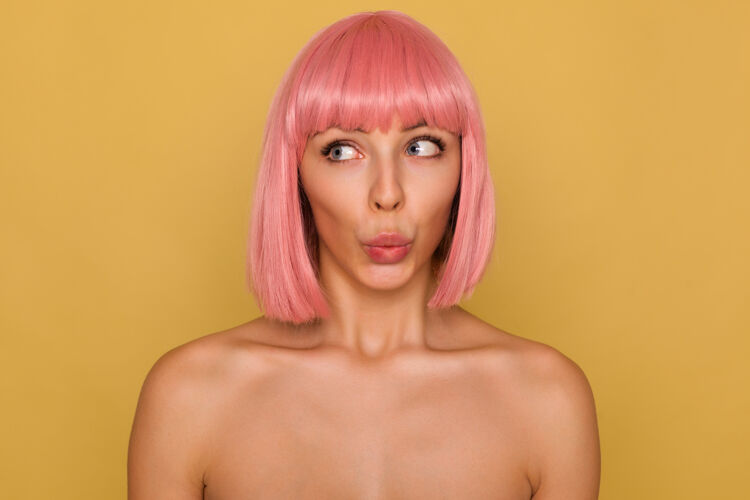 肖像激动的年轻漂亮女士的特写镜头 粉红色的短发叠着嘴唇 困惑地看着一边 赤裸着肩膀在芥末墙上摆姿势手头发女性