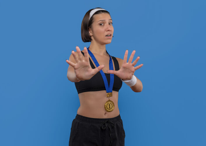 颈部一位身穿运动服 脖子上戴着金牌的年轻女子站在蓝色的墙壁上做着双手害怕的防守姿势运动员防御人物