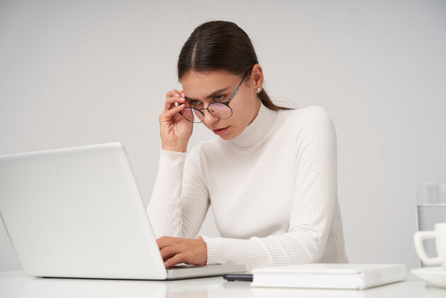 Poloneck严肃的年轻迷人的黑发女性 自然妆容 在办公室用笔记本电脑工作 穿着白色针织圆领衫和眼镜 在白色墙壁上摆姿势工作黑发发型